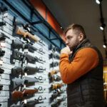 Assault Weapon BAN Backfires—Shelves Stripped Bare… Man contemplating firearms displayed on wall.