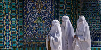 Three women in white traditional clothing, patterned wall background.