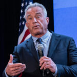 A man in a suit speaking into a microphone at a political event with an American flag in the background