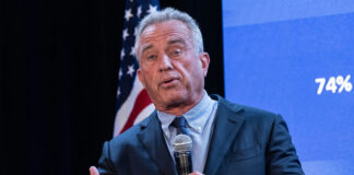 A man in a suit speaking into a microphone at a political event with an American flag in the background