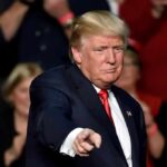 Wallet Rage Surges — Trump Takes The Hit man in suit pointing toward the camera at an event
