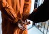 70-Year Hammer Hangs Over Trafficker Prisoner in orange jumpsuit handcuffed by officer