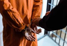 70-Year Hammer Hangs Over Trafficker Prisoner in orange jumpsuit handcuffed by officer