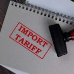 10% Wall Slams Imports—What Gets Hit Next? Notebook with import tariff stamp and red-handled rubber stamp