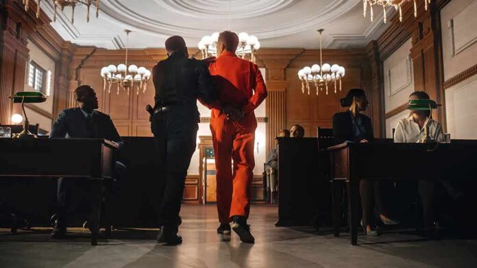 shutterstock_2056444886 Prisoner in orange jumpsuit escorted by officer in a courtroom