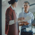 Workers Panic: Replaceable After One Click? Two colleagues discussing documents in a glass-walled office