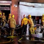Firefighters work outside a damaged warehouse with debris and hoses