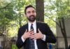 man in a suit speaking outdoors with hands on chest