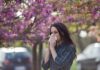 Early Wave Warning: Shot Now, Not Later Woman sneezing into tissue under blooming pink trees