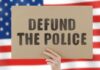 Tape Vs. Denial: El-Sayed Caught? Hand holding protest sign against blurred American flag