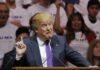 Donald Trump speaking at a rally, gesturing with one finger raised at a podium