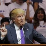 Donald Trump speaking at a rally, gesturing with one finger raised at a podium