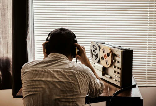 Person listening to tape recorder with headphones by window.
