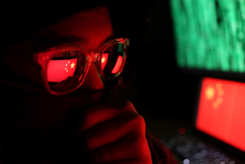 Person wearing glasses with a reflection of the Chinese flag, surrounded by computer screens